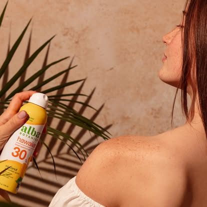 Femme appliquant une protection solaire sur son épaule, arrière-plan végétal.